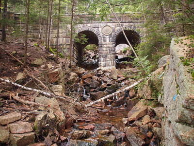 [This is a short but relatively tall double-arched stone bridge. It uses a variety of sizes of stones for its walls and center posts. The circular center stone is between the two arches. The brook which flows under it has many many large stones and not a lot of water flow.]